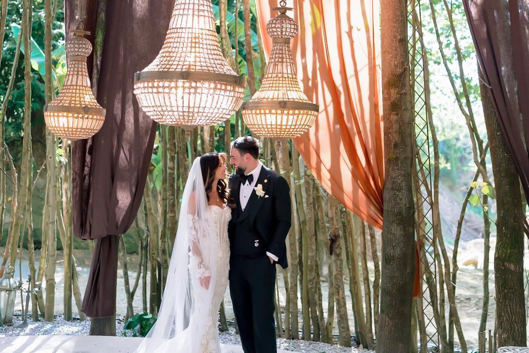 Bride and groom beneath chandeliers, with the groom wearing black tie tailoring