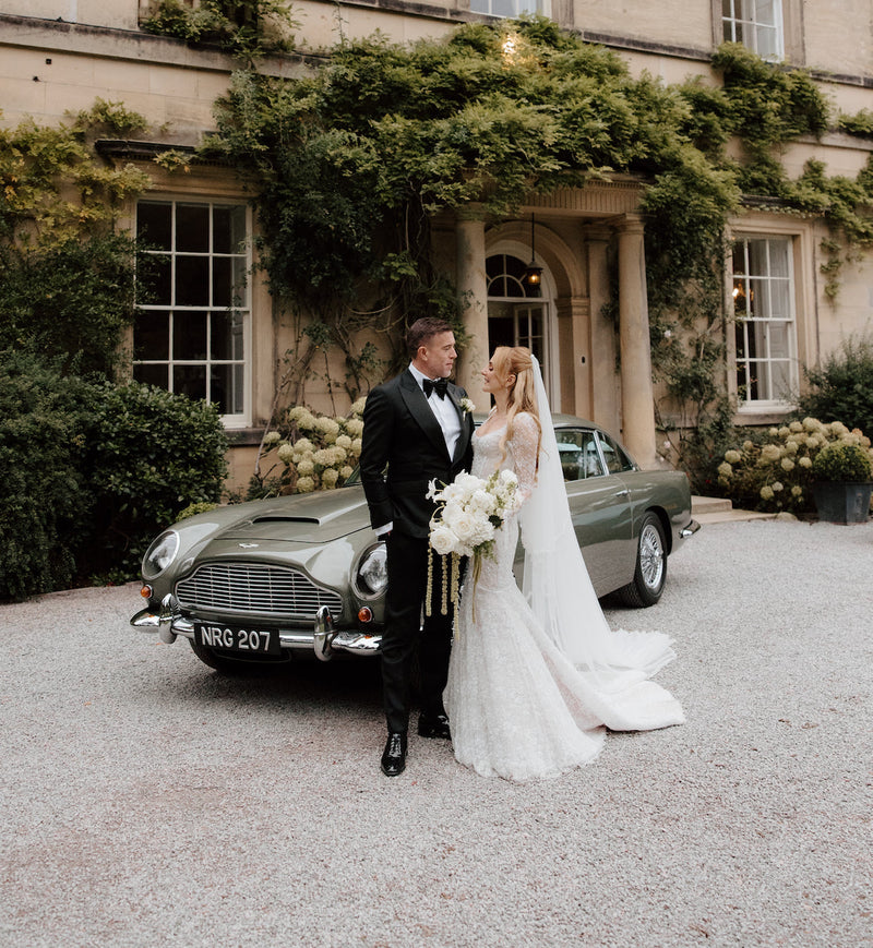 Bride and groom beside a classic Aston Martin outside a country house