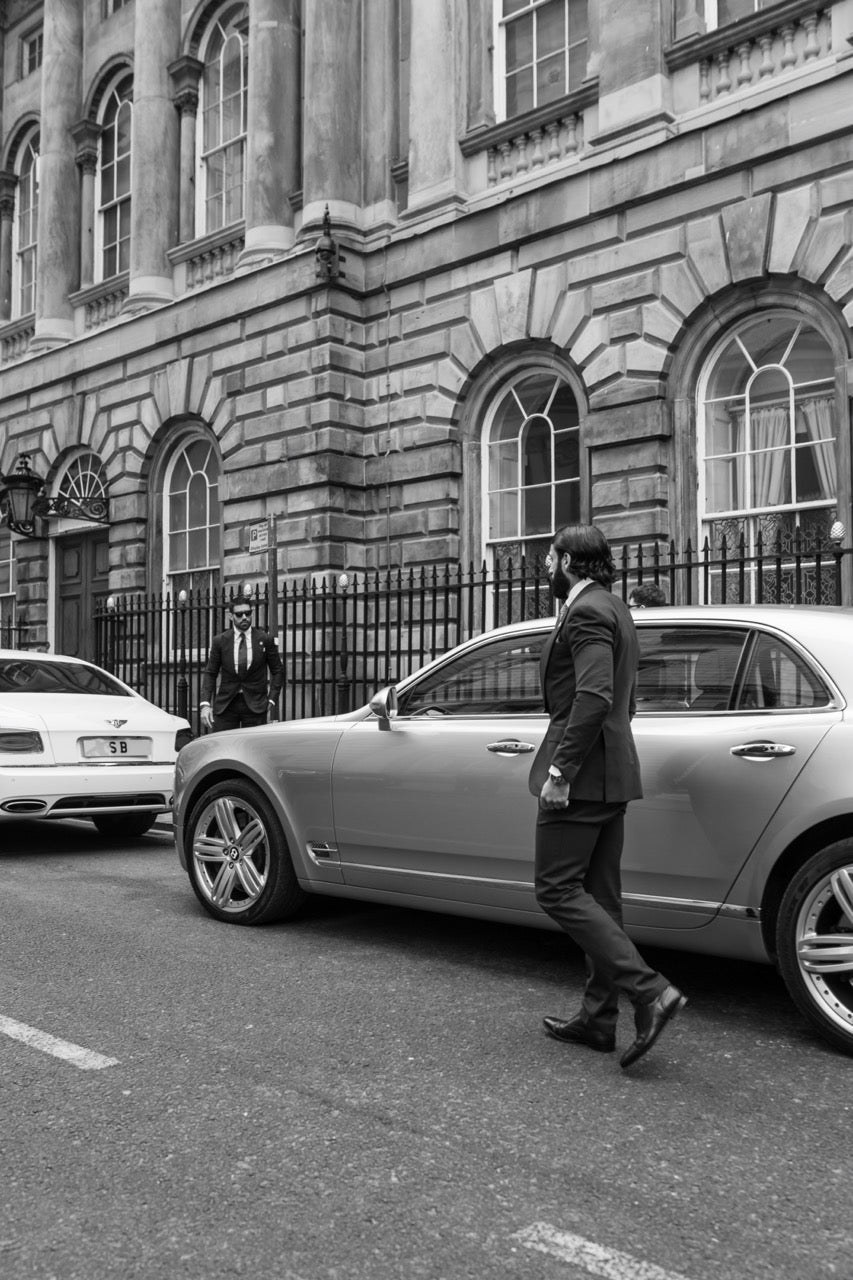 Man in a tailored suit walking beside luxury cars outside a city building